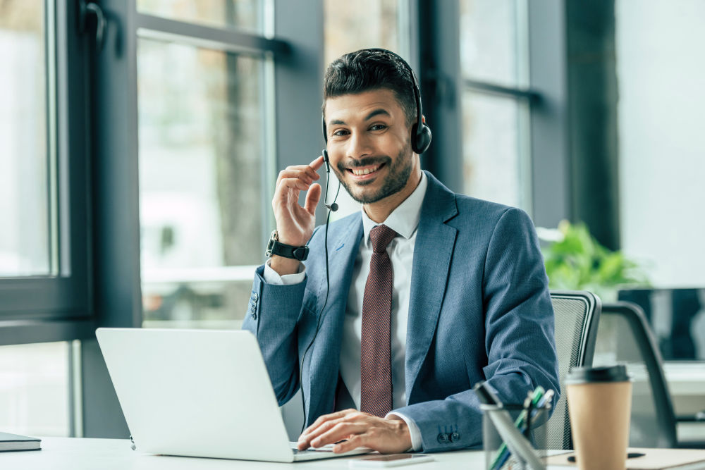 Cheerful call center operator looking at camera while sitting at workplace near laptop | How To Create An Effective Sales Call Script (Plus Free Template!) | sales script | sales phone script